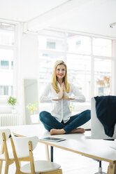 Smiling businesswoman sitting on desk in office practicing yoga - MOEF00229