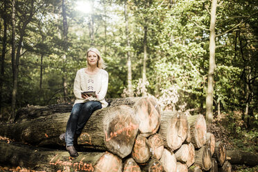 Portrait of woman sitting on stack of logs in the forest with tablet - MOEF00252