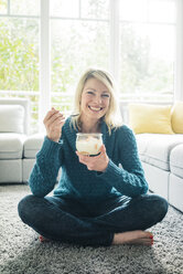 Porträt einer glücklichen Frau, die im Wohnzimmer einen Fruchtjoghurt isst - MOEF00279