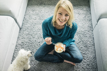 Porträt einer glücklichen Frau mit Hund, die im Wohnzimmer einen Fruchtjoghurt isst - MOEF00280