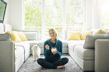 Porträt einer glücklichen Frau mit Hund, die im Wohnzimmer einen Fruchtjoghurt isst - MOEF00281