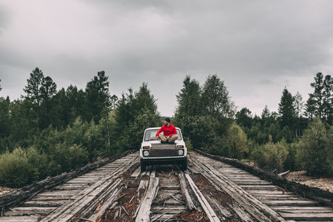 Man sitting on car bonnet on wooden lane in rural landscape stock photo