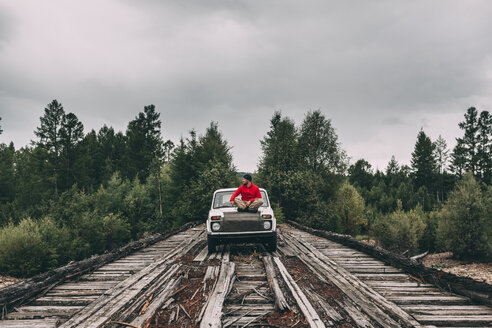 Man sitting on car bonnet on wooden lane in rural landscape - VPIF00235