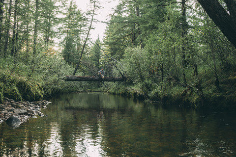 Mann sitzt auf einem Baumstamm über einem Fluss im Wald, lizenzfreies Stockfoto