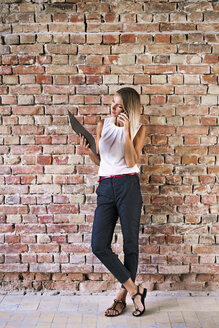 Geschäftsfrau, die ein Klemmbrett hält und ein Mobiltelefon an einer Backsteinmauer im Büro benutzt - HAPF02348