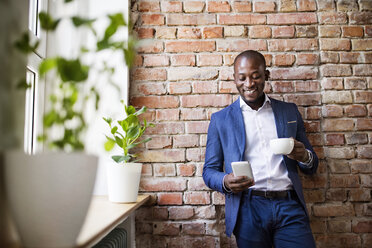 Smiling businessman using cell phone at brick wall by the window - HAPF02360