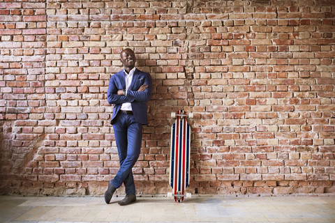 Lächelnder Geschäftsmann mit Longboard an Backsteinmauer, lizenzfreies Stockfoto