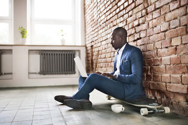 Businessman sitting on longboard at brick wall using laptop - HAPF02375