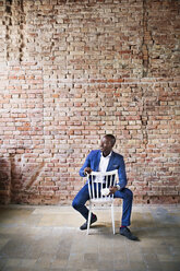 Smiling businessman with cup of coffee sitting on chair at brick wall - HAPF02376