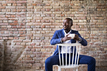 Lächelnder Geschäftsmann mit einer Tasse Kaffee auf einem Stuhl an einer Backsteinmauer sitzend - HAPF02377