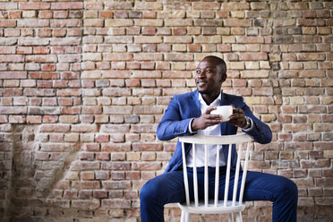 Smiling businessman with cup of coffee sitting on chair at brick wall - HAPF02377