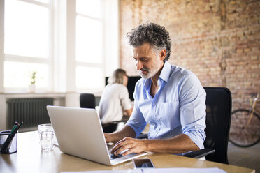 Businessman using laptop in office with colleague in background - HAPF02421