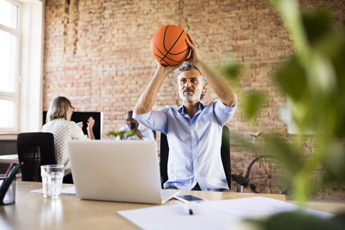 Businessman playing basketball in office with colleagues in background - HAPF02422