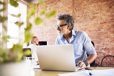 Businessman using laptop in office with colleague in background - HAPF02423