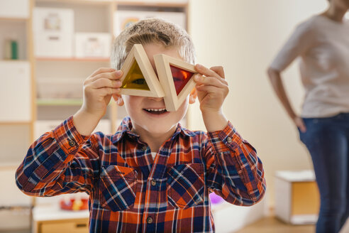 Boy holding wooden and colorful blocks before his eyes in kindergarten - MFF04088