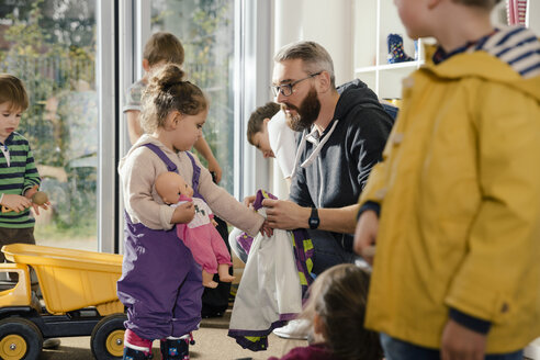 Pre-school teacher helping girl putting on rainwear in kindergarten - MFF04106