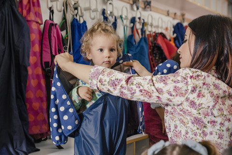 Vorschullehrerin hilft Mädchen beim Anziehen der Matschhose im Kindergarten, lizenzfreies Stockfoto