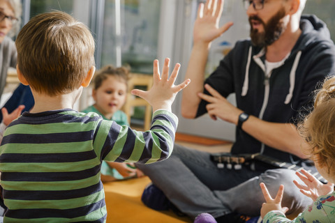 Kind hebt die Hand beim Singen mit anderen und der Lehrerin im Kindergarten, lizenzfreies Stockfoto