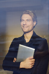 Portrait of smiling young businessman with tablet behind windowpane - PNEF00246