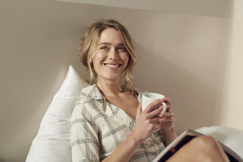 Porträt einer glücklichen Frau auf dem Bett sitzend mit Kaffeetasse und Zeitschrift - PNEF00262