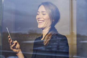 Portrait of laughing businesswoman behind windowpane looking at cell phone - PNEF00282