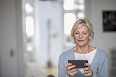 Porträt einer älteren Frau zu Hause, die auf ihr Mobiltelefon schaut - FMKF04611