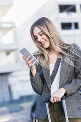 Happy young woman with rolling suitcase checking cell phone - UUF12221