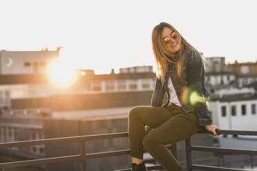 Young woman sitting on railing in the city at sunset - UUF12235