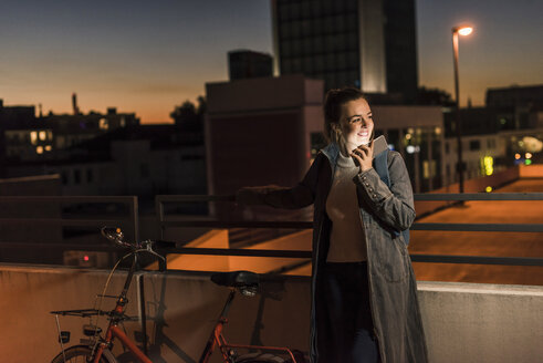 Lächelnde junge Frau in der Stadt beim Telefonieren am Abend - UUF12249
