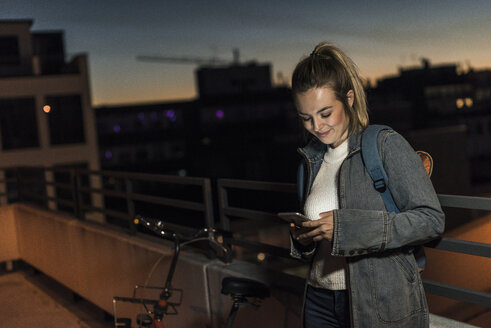 Lächelnde junge Frau in der Stadt, die am Abend ihr Handy überprüft - UUF12250