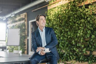 Laughing businessman in green office with cup of coffee - JOSF01815