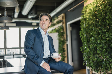 Smiling businessman in green office with cup of coffee - JOSF01817