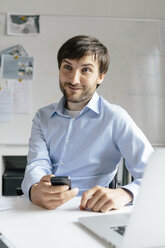 Smiling businessman with cell phone and laptop at desk in office - BMF00846