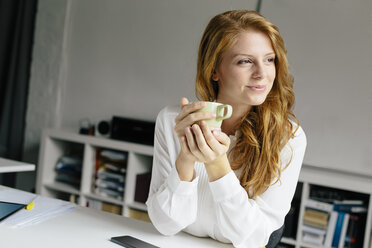 Lächelnde junge Frau mit einer Tasse Kaffee am Schreibtisch im Büro - BMF00864