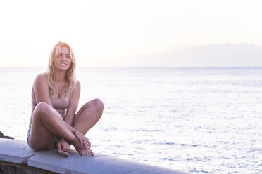 Portrait of smiling blond woman sitting on a wall in front of the sea - SIPF01853