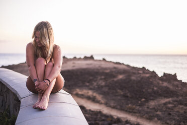 Spanien, Teneriffa, lachende blonde Frau sitzt bei Sonnenuntergang auf einer Mauer am Meer - SIPF01855