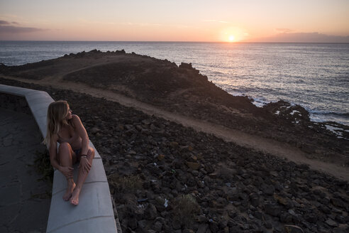 Spanien, Teneriffa, junge Frau sitzt auf einer Mauer und genießt den Sonnenuntergang am Meer - SIPF01859