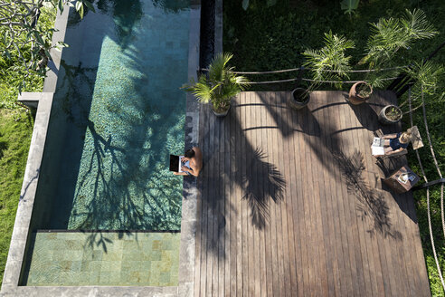 Mature man sitting at the poolside, using laptop, while daughter is reading - SBOF00855