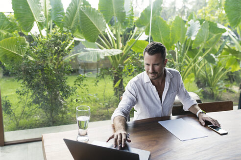 Mature man sitting at table in front of lush garden, using laptop - SBOF00869
