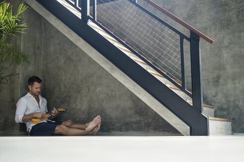 Mature man sitting under staircase, playing the ukulele - SBOF00870