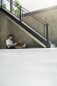 Mature man sitting under staircase, reading a book - SBOF00872