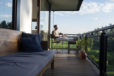 Mann sitzt auf dem Balkon eines modernen Hauses und schaut in die Landschaft, er hält einen Laptop - SBOF00875