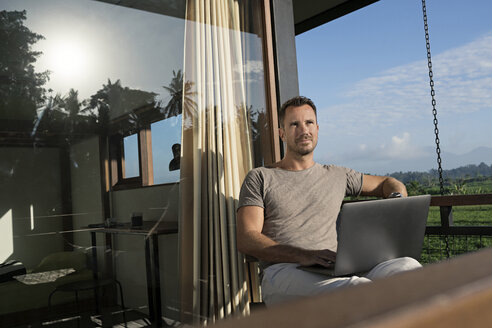 Man sitting on balcony of modern house looking landscape, holding laptop - SBOF00876