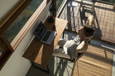 Man leaning back and taking a break from working at laptop on desk in house - SBOF00879