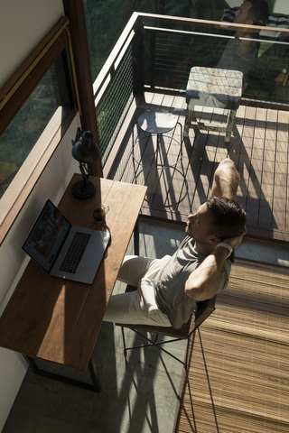 Mann lehnt sich zurück und macht eine Pause von der Arbeit am Laptop auf dem Schreibtisch im Haus, lizenzfreies Stockfoto