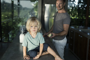 Sohn sitzt auf dem Tresen in der Küche, während der Vater das Frühstück vorbereitet - SBOF00887