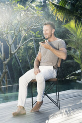 Mature man sitting barefoot by the poolside, talking on the phone - SBOF00893