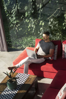 Reifer Mann auf roter Couch sitzend, mit Laptop, arbeitend - SBOF00894