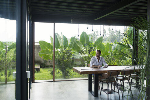 Mature man sitting at table in front of lush garden, writing - SBOF00906