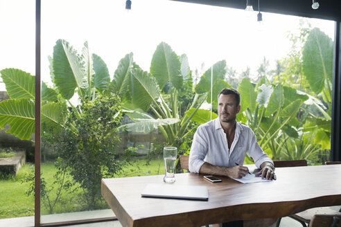 Mature man sitting at table in front of lush garden, writing - SBOF00907